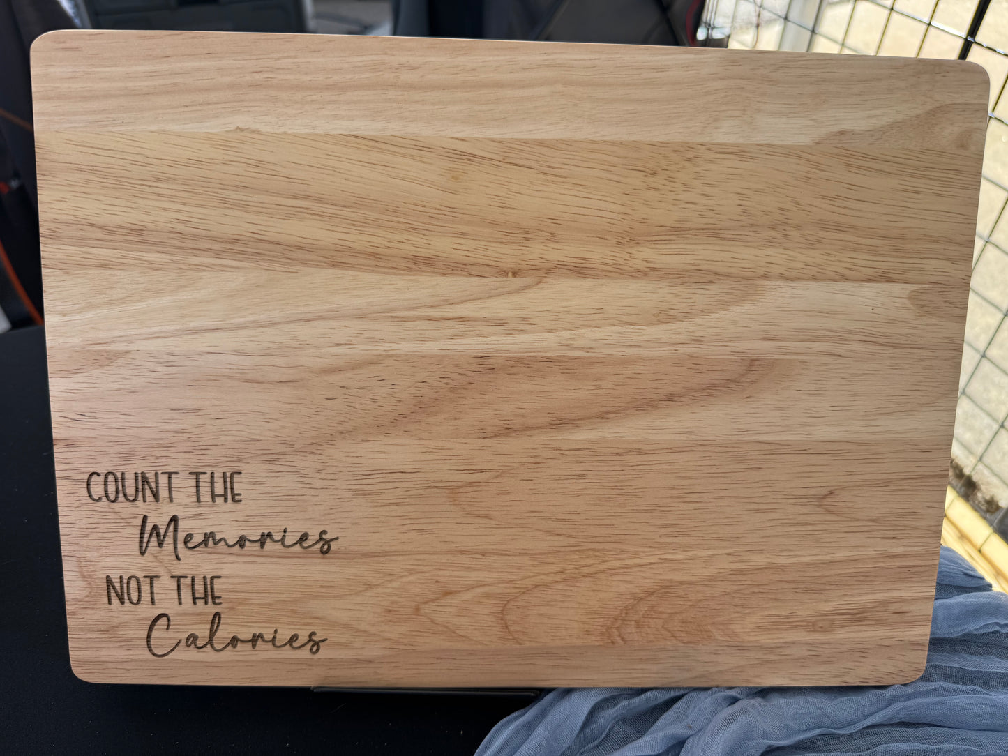 Laser-engraved rubber wood cutting board with the quote “Count the Memories Not the Calories” in a mix of block and script fonts, displayed upright on a table with a mesh cloth background.