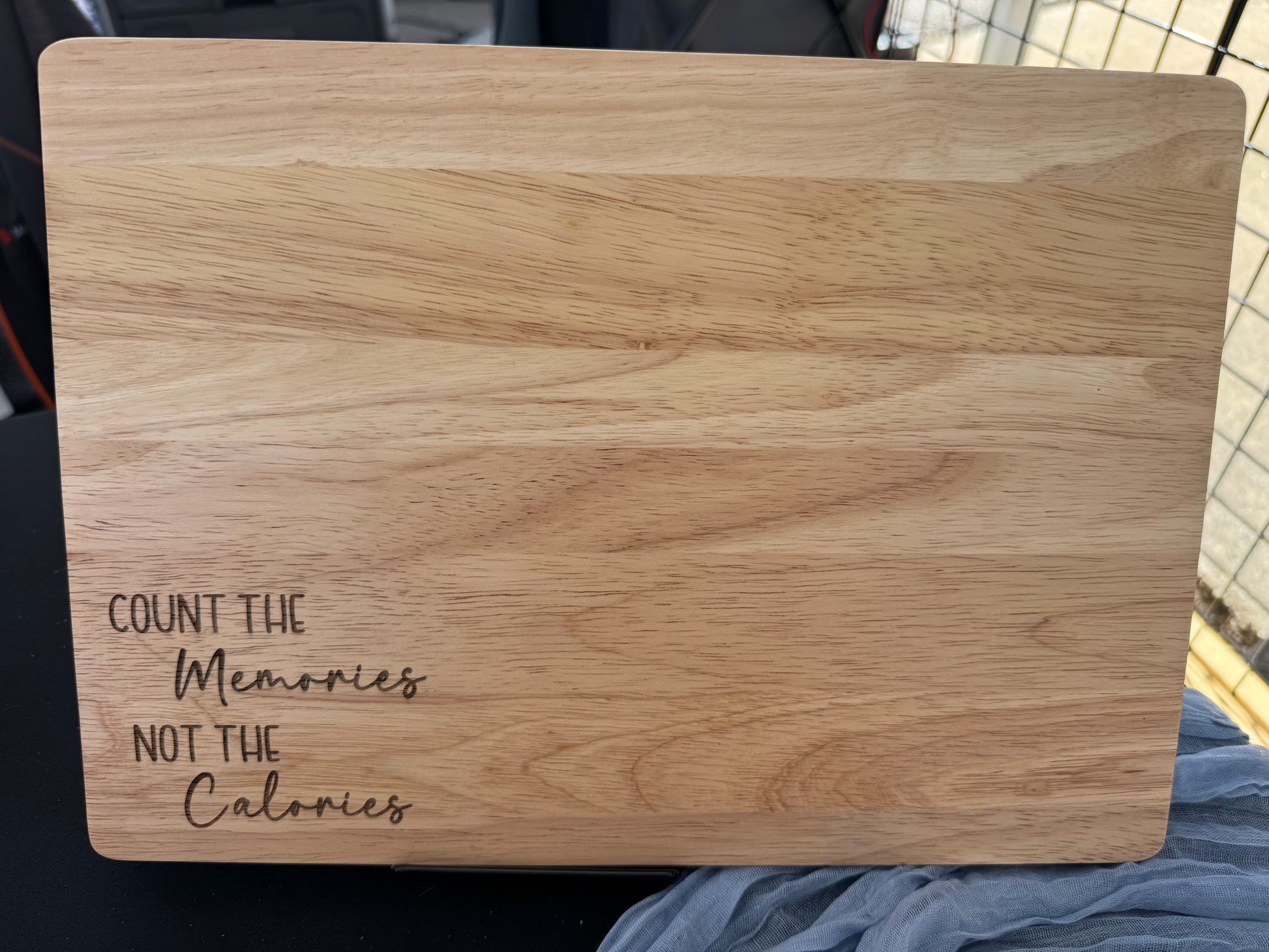 Laser-engraved rubber wood cutting board with the quote “Count the Memories Not the Calories” in a mix of block and script fonts, displayed upright on a table with a mesh cloth background.