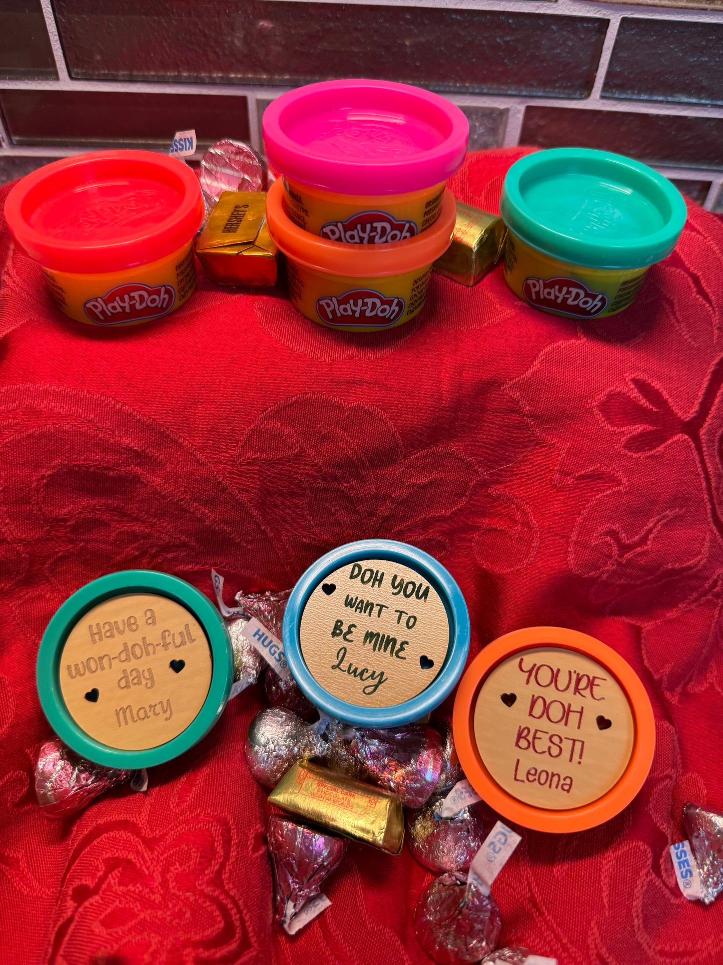 Close-up view of mini ‘Doh containers featuring personalized engraved wooden lids with custom Valentine’s Day phrases, set on a festive red background.