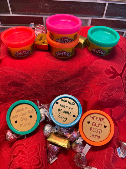 Close-up view of mini ‘Doh containers featuring personalized engraved wooden lids with custom Valentine’s Day phrases, set on a festive red background.