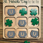 Close-up of a St. Patrick’s Day-themed tic-tac-toe board with green shamrocks and gray pots of gold as player pieces. The engraved title reads ‘St. Patrick’s Day Tic-Tac-Toe,’ displayed on a rustic wooden background.