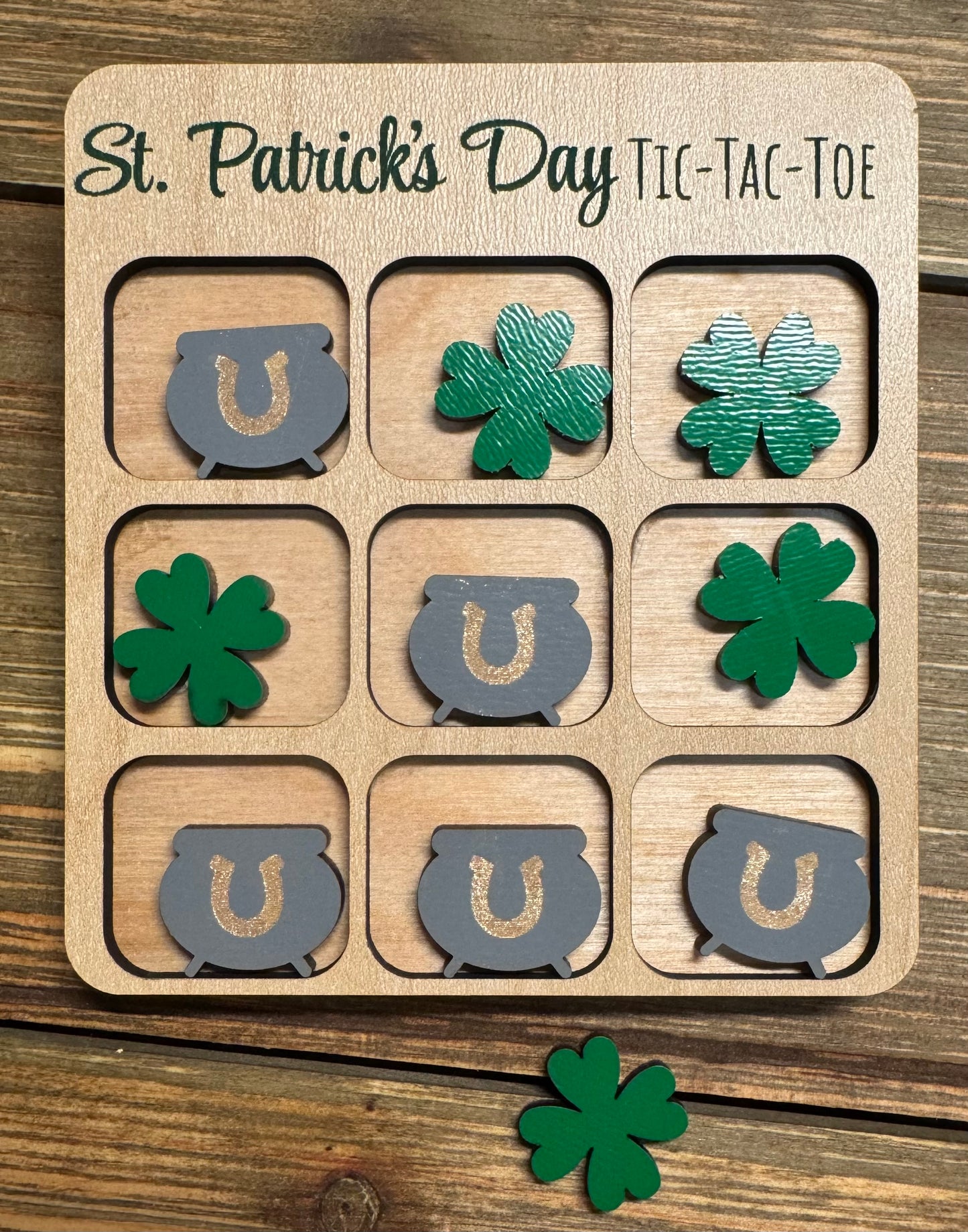 Close-up of a St. Patrick’s Day-themed tic-tac-toe board with green shamrocks and gray pots of gold as player pieces. The engraved title reads ‘St. Patrick’s Day Tic-Tac-Toe,’ displayed on a rustic wooden background.