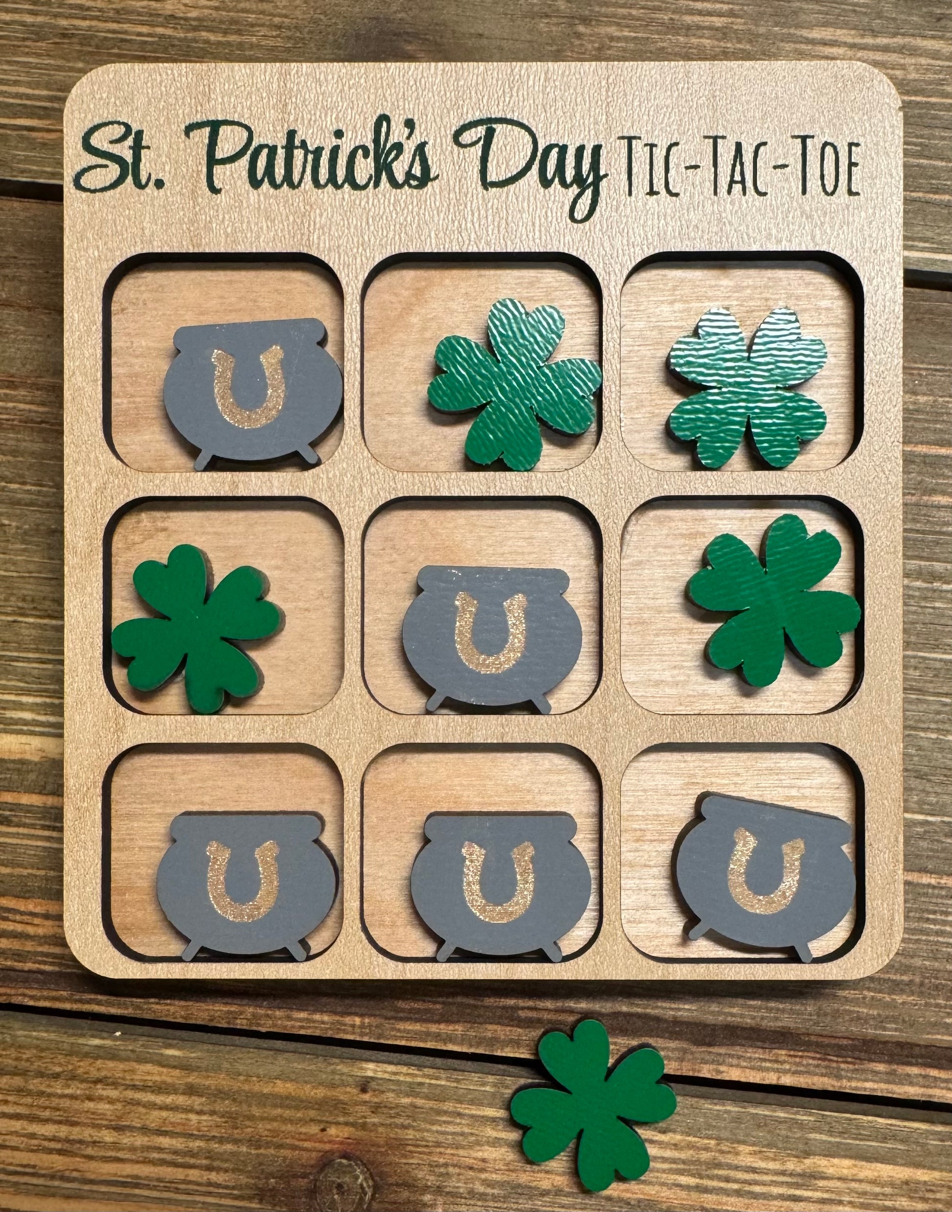 Close-up of a St. Patrick’s Day-themed tic-tac-toe board with green shamrocks and gray pots of gold as player pieces. The engraved title reads ‘St. Patrick’s Day Tic-Tac-Toe,’ displayed on a rustic wooden background.