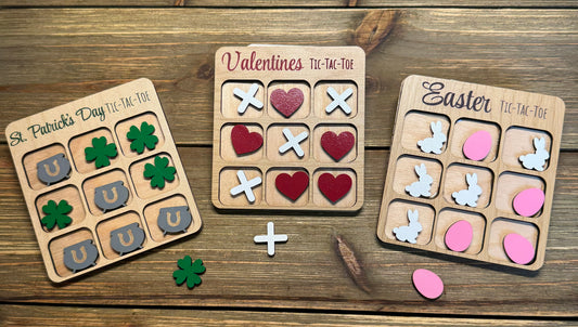 Set of three holiday-themed wooden tic-tac-toe boards displayed on a rustic wood surface. Designs include Valentine’s Day with hearts and X’s, St. Patrick’s Day with shamrocks and pots of gold, and Easter with bunnies and eggs. Perfect for holiday gifts, party favors, and stocking stuffers.