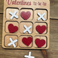 Close-up of a Valentine’s Day-themed tic-tac-toe board with red hearts and white X’s as player pieces. The engraved title reads ‘Valentines Tic-Tac-Toe,’ set on a rustic wooden surface.