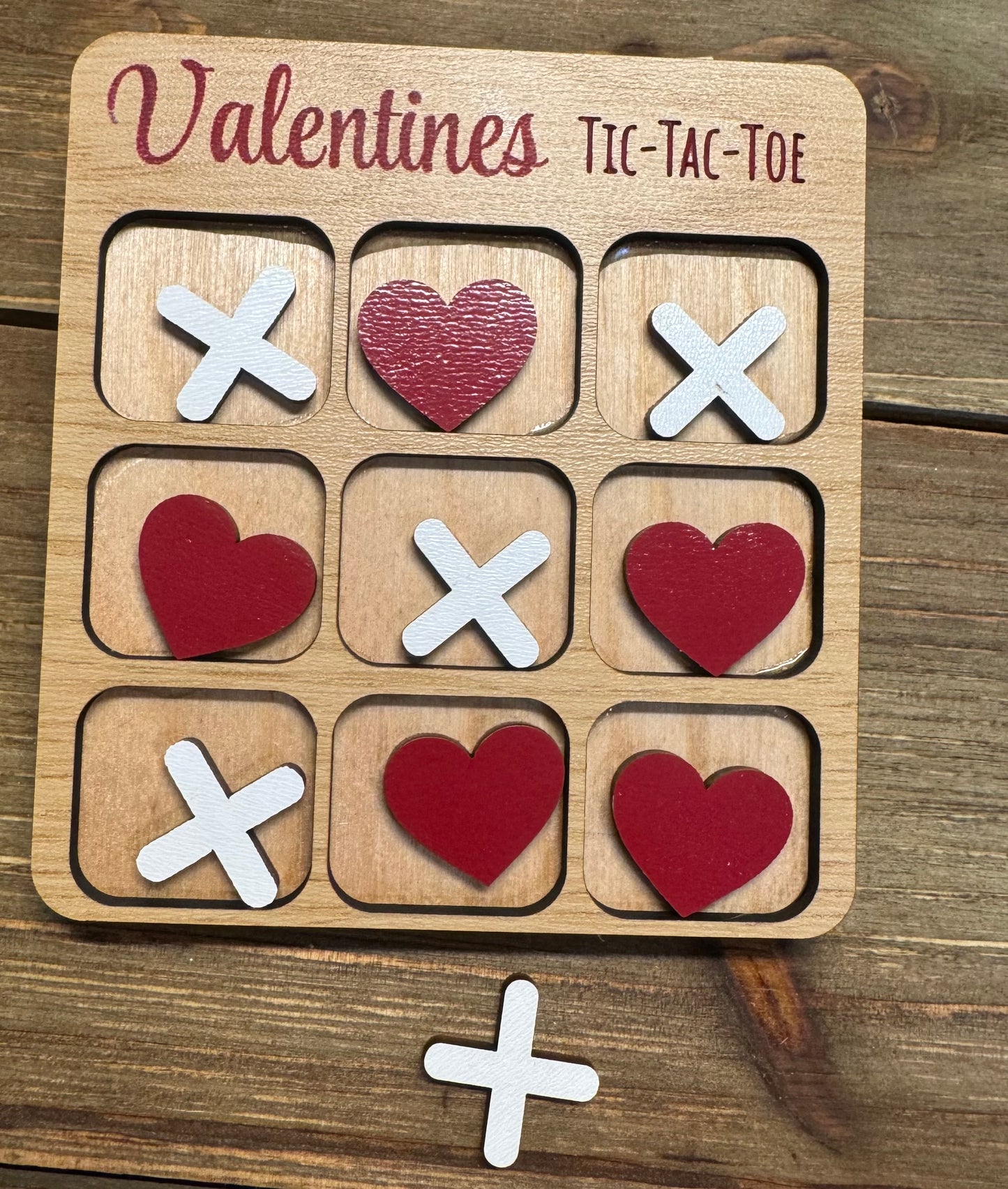 Close-up of a Valentine’s Day-themed tic-tac-toe board with red hearts and white X’s as player pieces. The engraved title reads ‘Valentines Tic-Tac-Toe,’ set on a rustic wooden surface.