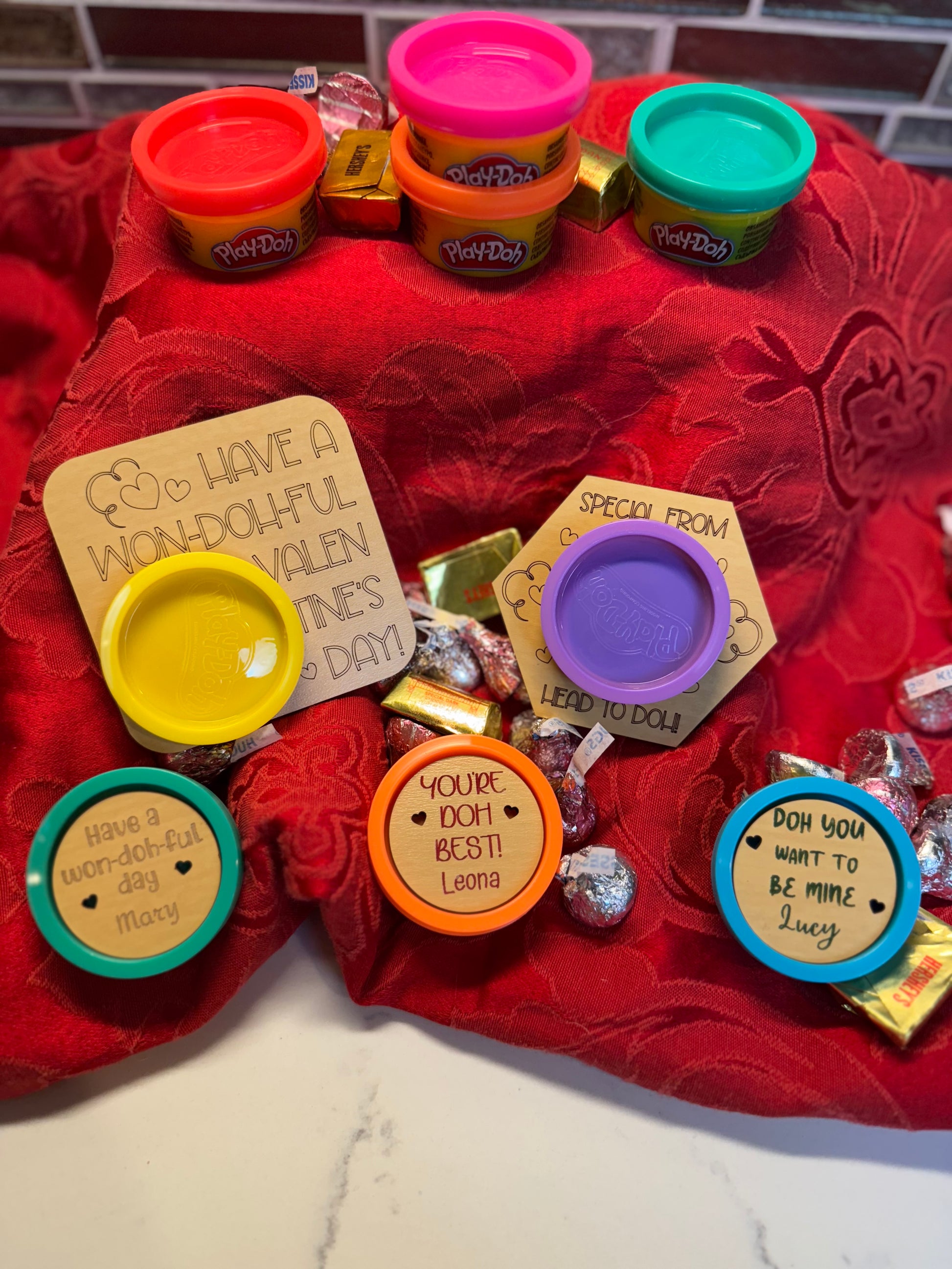 Set of 8 colorful mini Play-Doh containers with personalized engraved wooden lids, displayed on a red decorative cloth. Perfect for Valentine’s Day gifts, party favors, or classroom exchanges.