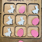 Close-up of an Easter-themed tic-tac-toe board featuring pink eggs and white bunnies as player pieces. The engraved title reads ‘Easter Tic-Tac-Toe,’ displayed on a rustic wood background.