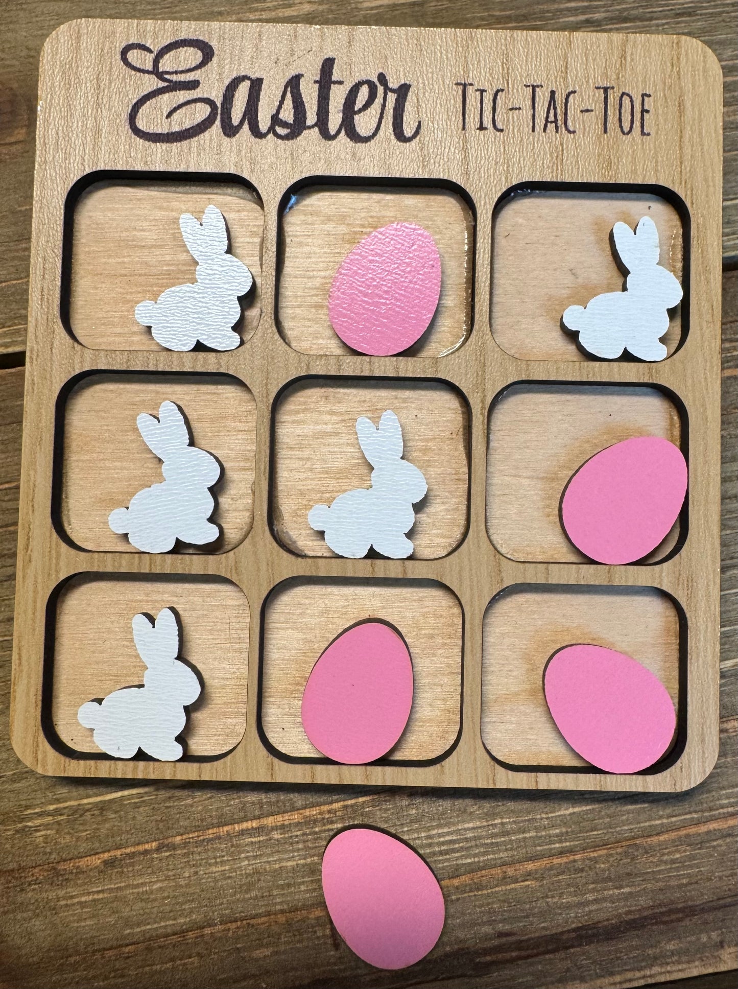 Close-up of an Easter-themed tic-tac-toe board featuring pink eggs and white bunnies as player pieces. The engraved title reads ‘Easter Tic-Tac-Toe,’ displayed on a rustic wood background.