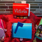 A display of four colorful mini tin mailboxes in red, pink, blue, and green, set on a red decorative cloth. Each mailbox is filled with assorted chocolates and candies, with the red mailbox personalized with the name ‘Victoria’ in white script. The mailboxes have hinged front openings and flags, showcasing their Valentine’s Day theme.