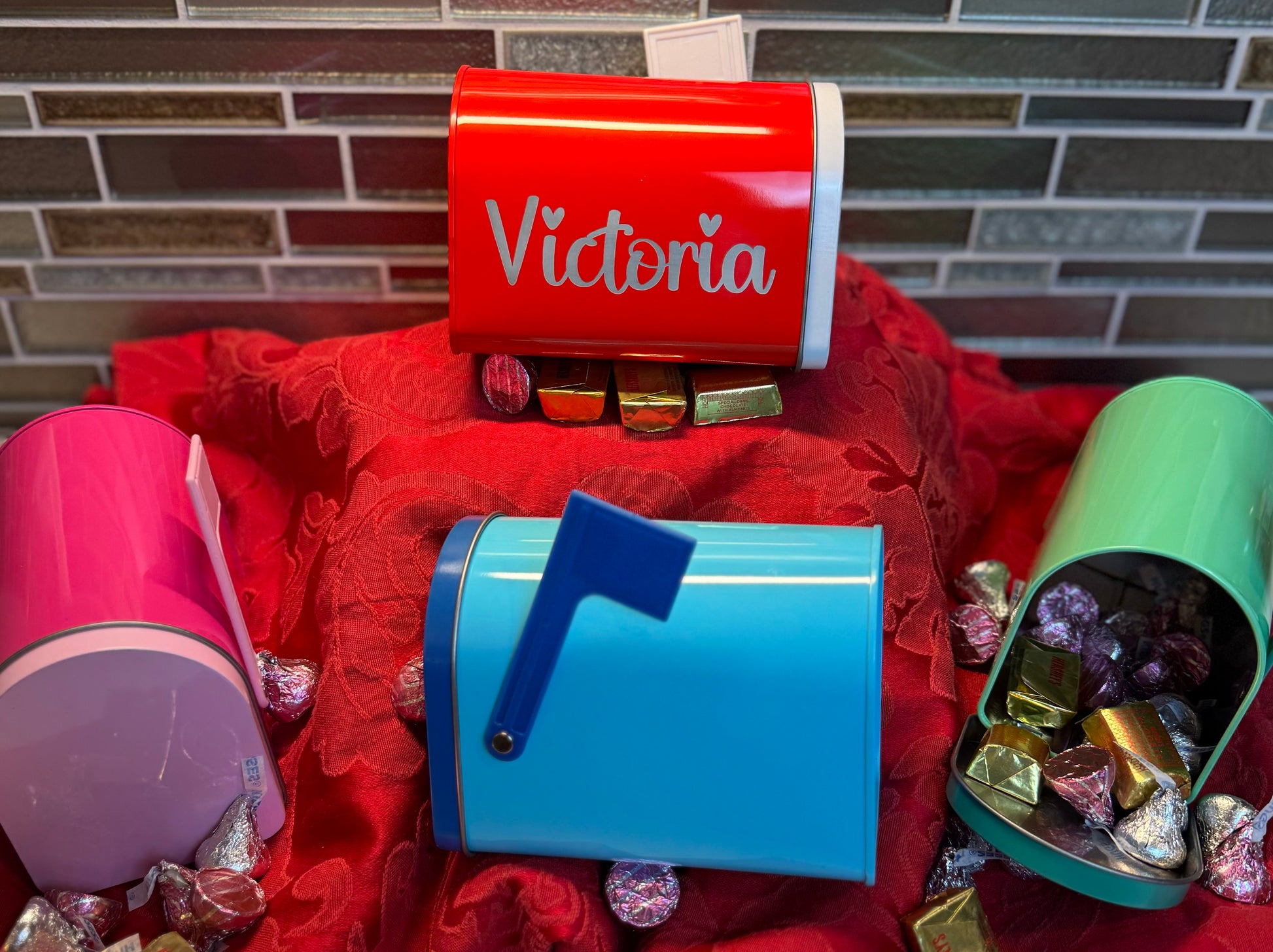 A display of four colorful mini tin mailboxes in red, pink, blue, and green, set on a red decorative cloth. Each mailbox is filled with assorted chocolates and candies, with the red mailbox personalized with the name ‘Victoria’ in white script. The mailboxes have hinged front openings and flags, showcasing their Valentine’s Day theme.