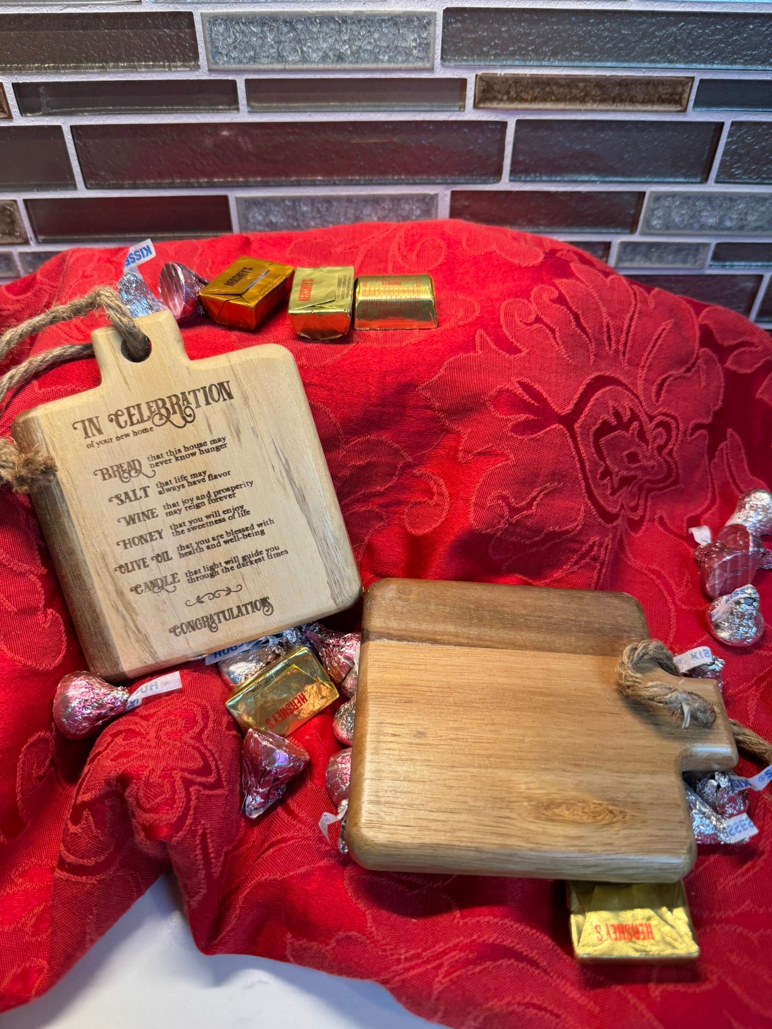 Rustic mini cutting board set displayed on a red fabric backdrop, featuring an engraved blessing and a plain wooden board, surrounded by chocolates.