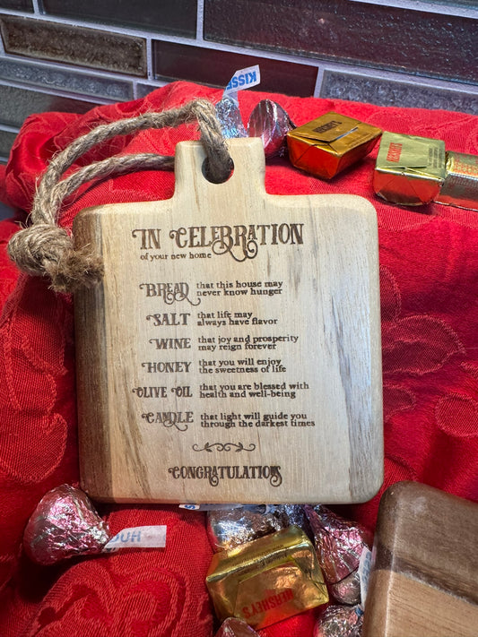 Close-up of a rustic mini cutting board with jute rope, engraved with a housewarming blessing celebrating bread, salt, wine, honey, olive oil, and a candle for prosperity and joy.