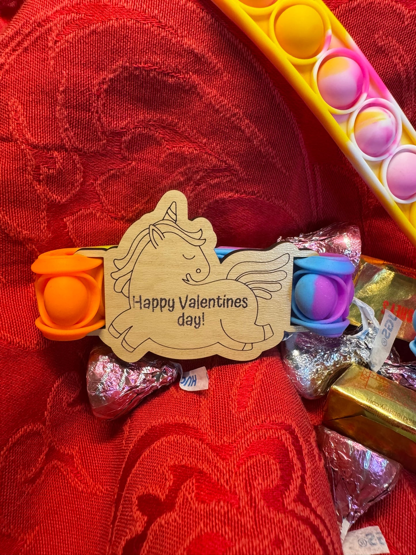 Colorful Pop It fidget toy with a laser-engraved wooden unicorn tag reading 'Happy Valentines Day!' displayed on a red textured background with chocolate candies