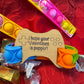 Pop It fidget toy in vibrant rainbow colors, paired with a wooden tag shaped like a gaming controller engraved with the message 'I hope your Valentine’s is poppin’!' surrounded by candy on a red background.