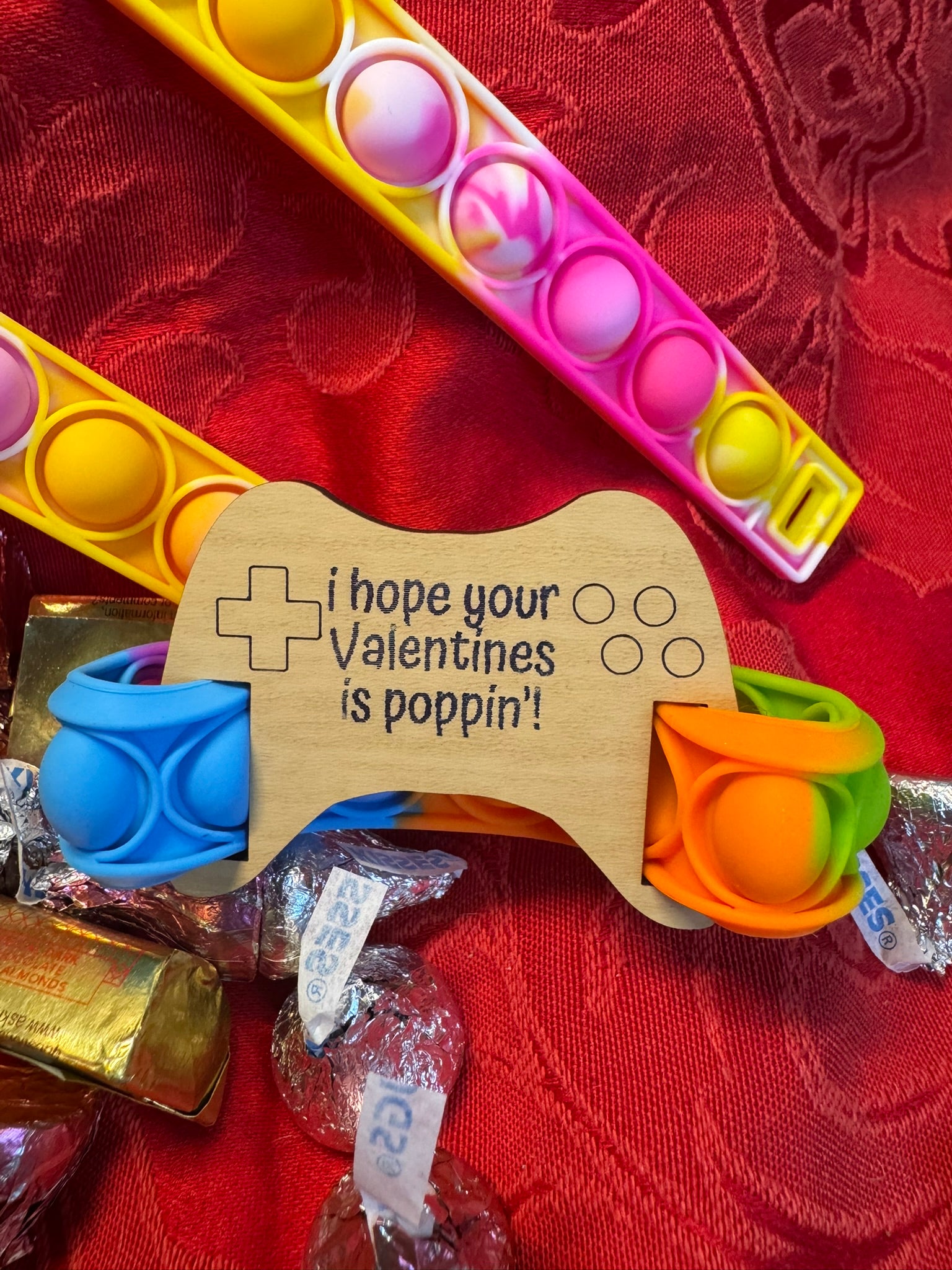 Pop It fidget toy in vibrant rainbow colors, paired with a wooden tag shaped like a gaming controller engraved with the message 'I hope your Valentine’s is poppin’!' surrounded by candy on a red background.