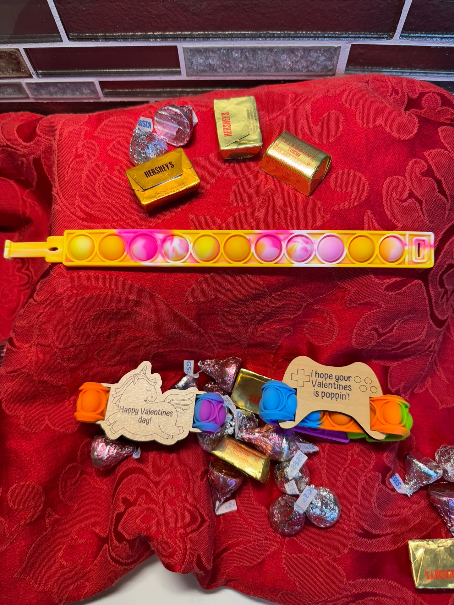 Valentine’s gift set featuring colorful Pop It fidget toys, laser-engraved wooden tags with festive messages, and decorative chocolates, arranged on a red fabric backdrop.