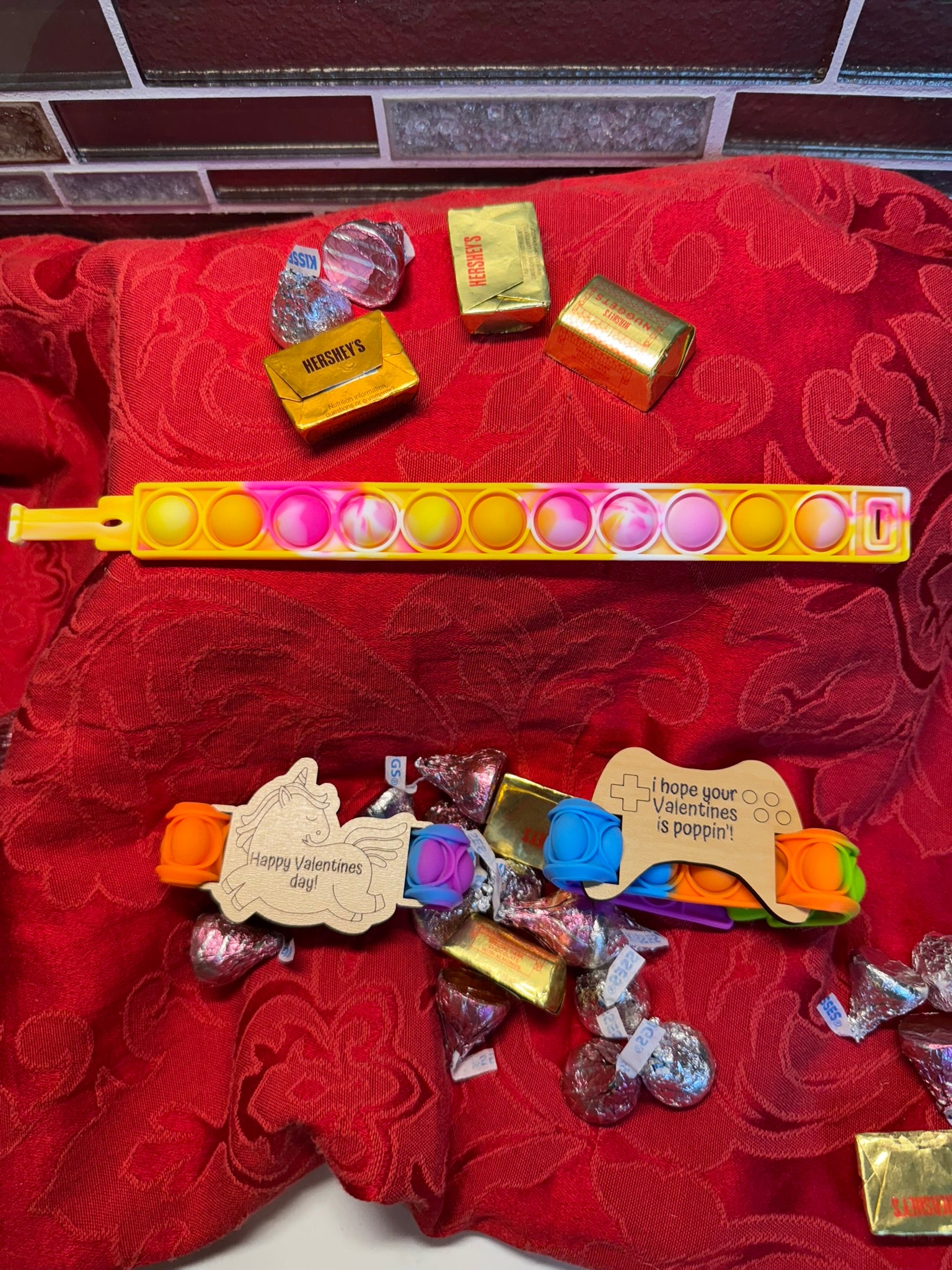 Valentine’s gift set featuring colorful Pop It fidget toys, laser-engraved wooden tags with festive messages, and decorative chocolates, arranged on a red fabric backdrop.