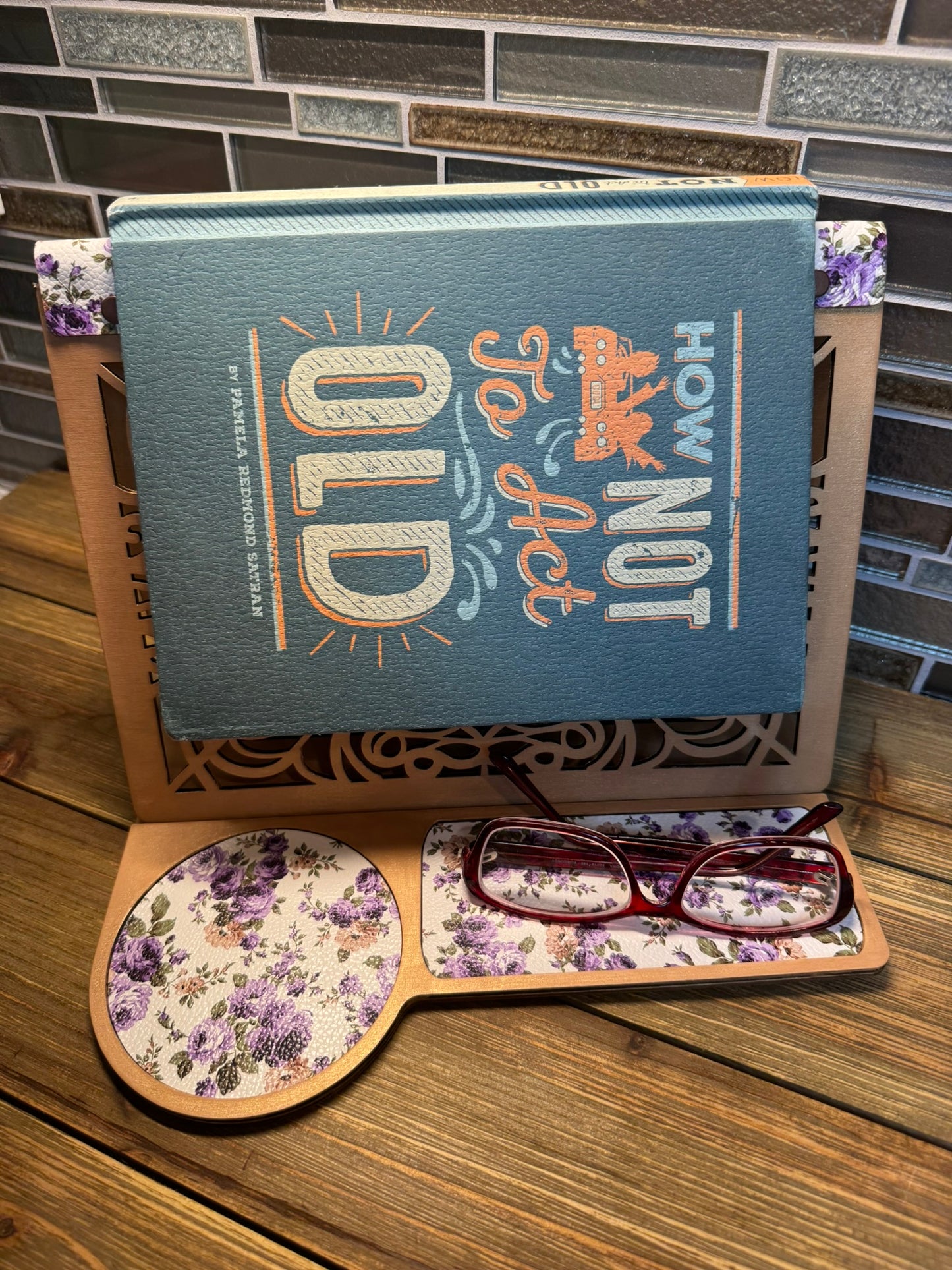 Handcrafted wooden book stand with a laser-cut design, holding a sample book featuring a purple floral pattern on the base and glasses resting on the side.