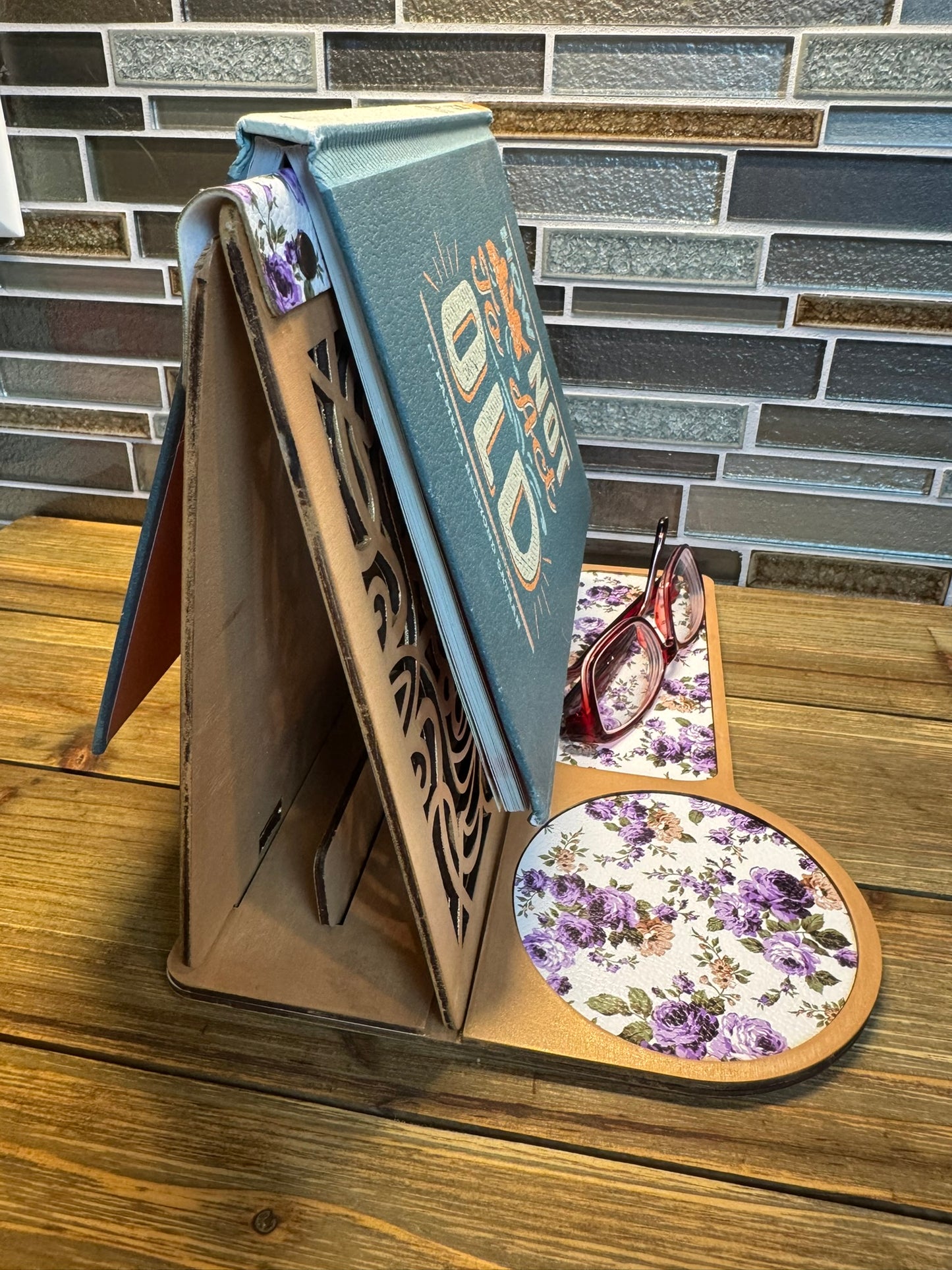 Handcrafted wooden book stand with a laser-cut design, holding a sample book featuring a purple floral pattern on the base and glasses resting on the side.