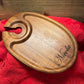 Oval-shaped wooden wine glass holder with a custom engraving reading 'Nicola,' displayed on a red fabric backdrop.