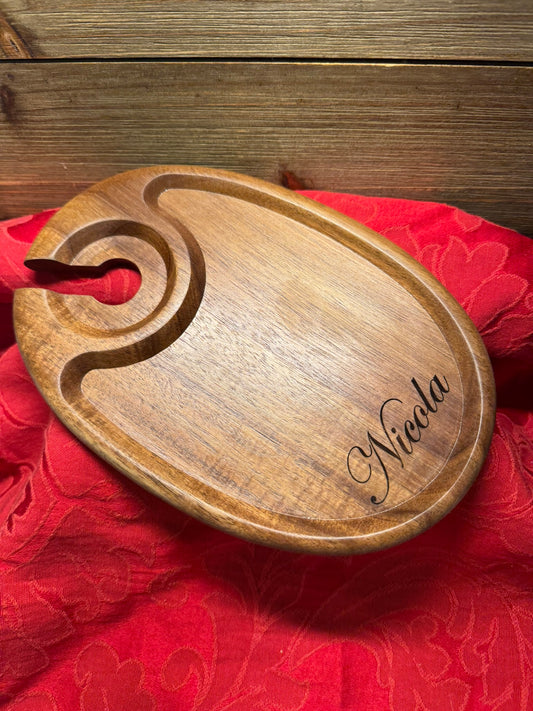 Oval-shaped wooden wine glass holder with a custom engraving reading 'Nicola,' displayed on a red fabric backdrop.