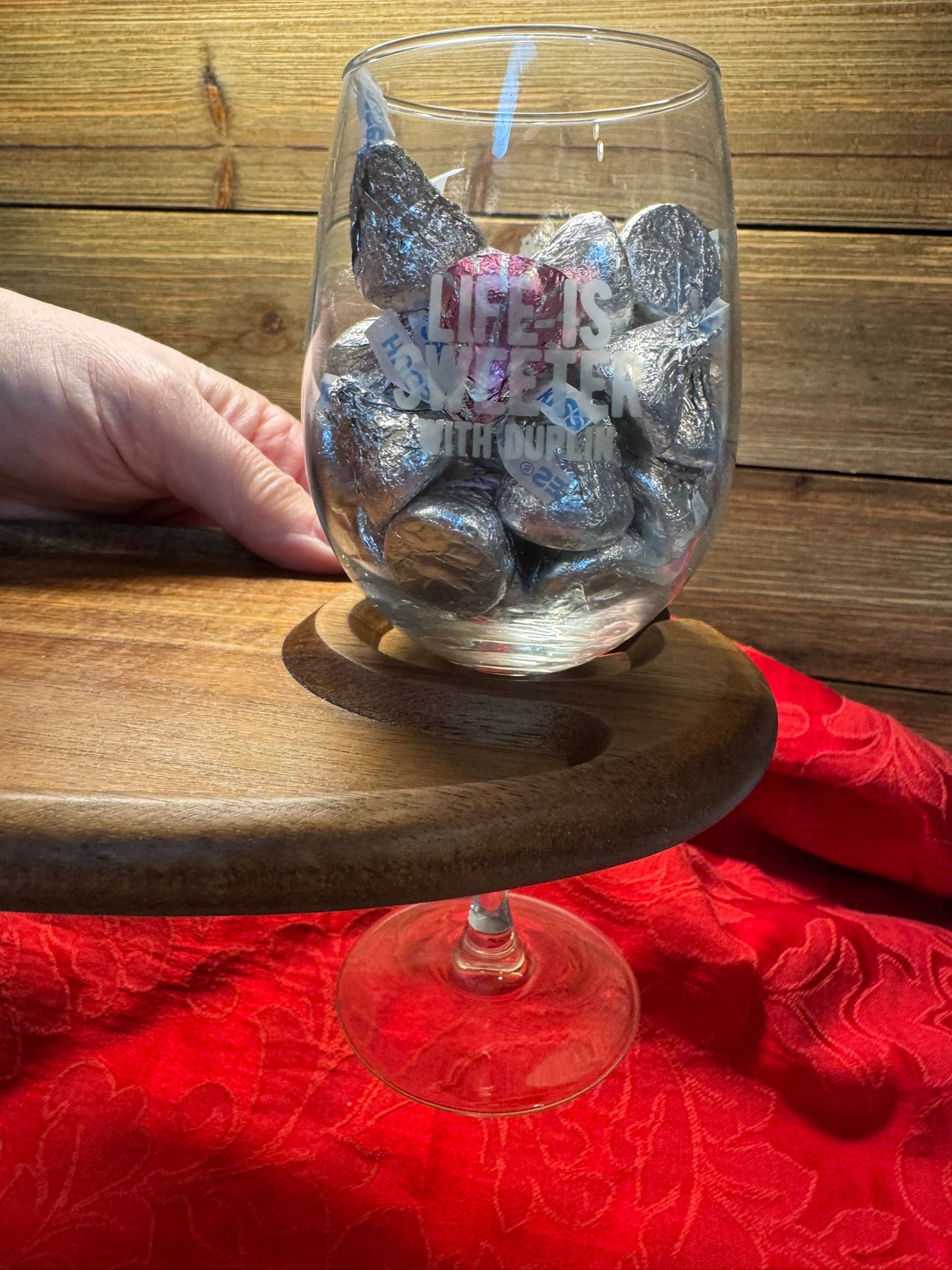 Close-up of a wooden wine glass holder in use, securely holding a stemmed glass filled with chocolates, with the engraving 'Enjoy' visible.