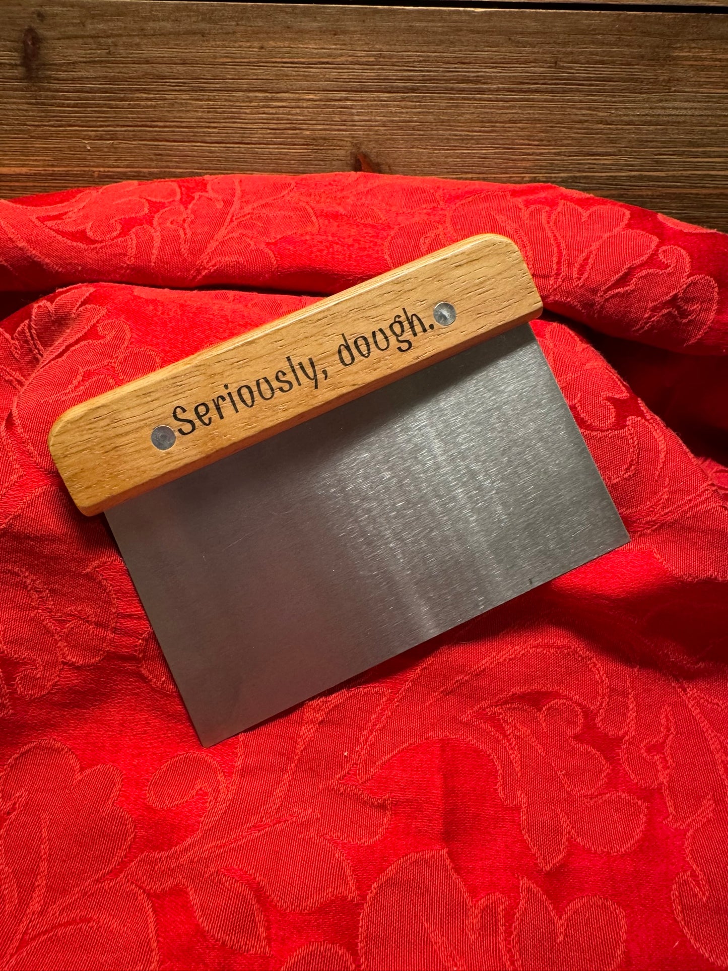 Bench scraper with a wooden handle engraved with the phrase 'Seriously, Dough,' displayed on a red textured fabric backdrop.