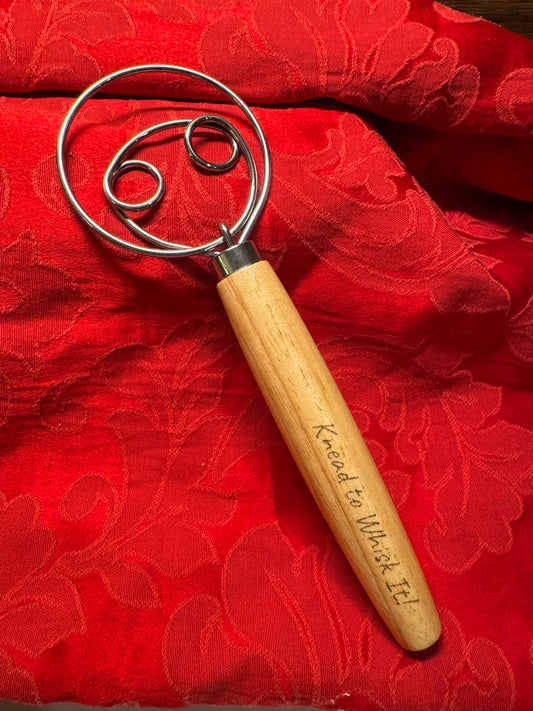 Close-up of a Danish dough whisk with an engraved wooden handle that reads 'Knead to Whisk It,' displayed on a red textured fabric backdrop.