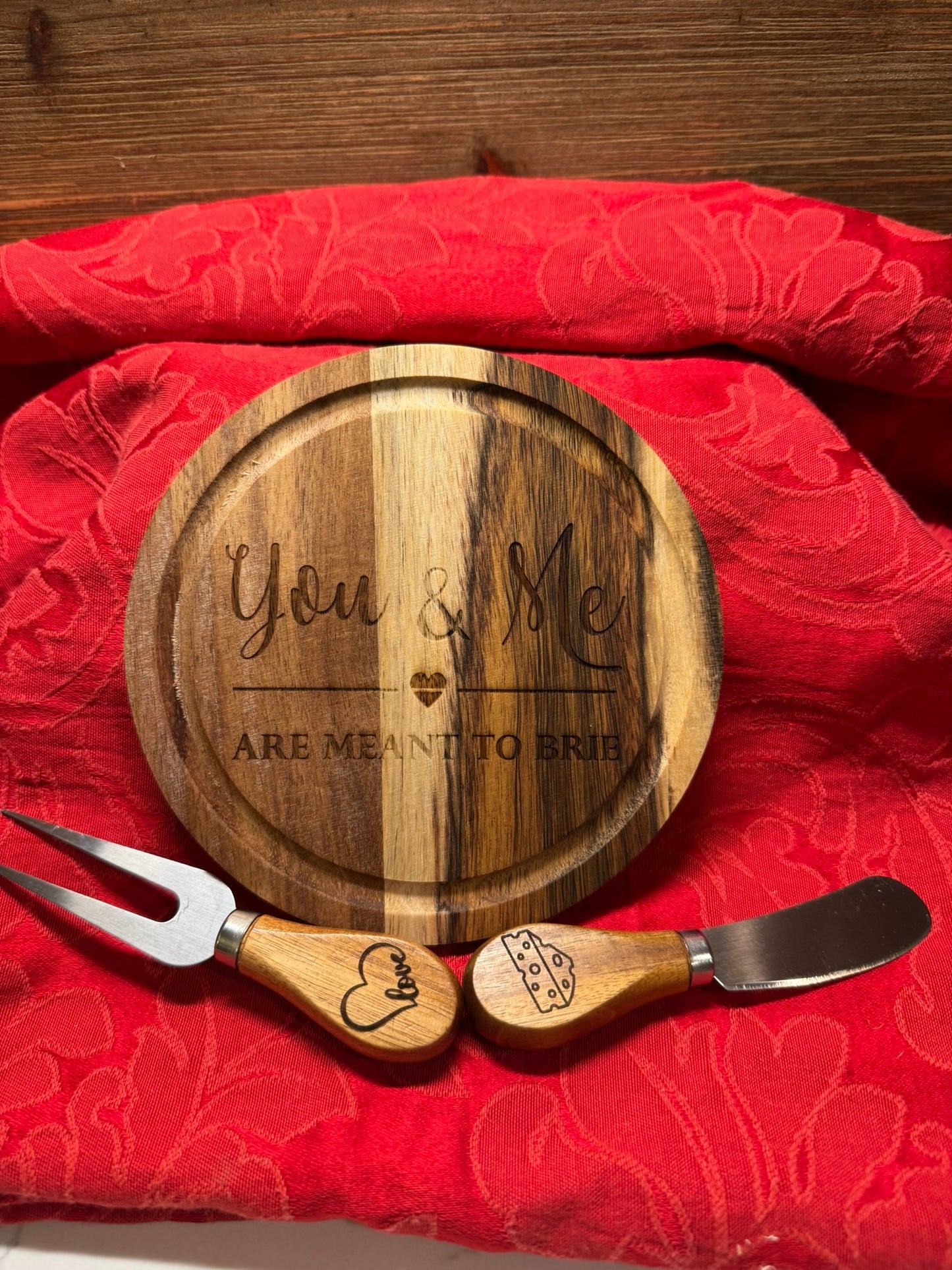 Engraved wooden cheese board reading 'You & Me Are Meant to Brie' with a matching cheese fork and spreader featuring heart and cheese engravings, displayed on a red fabric background.