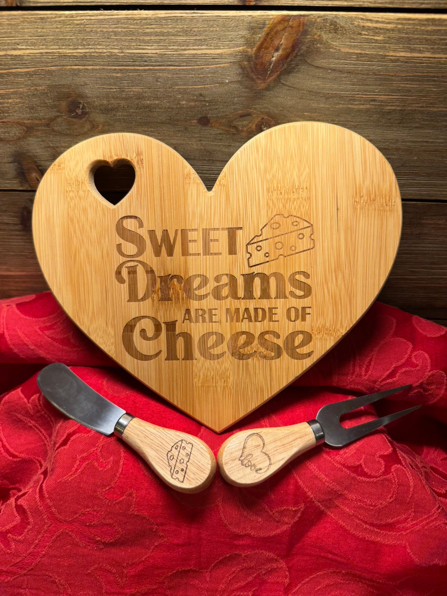 Heart-shaped wooden cheese board engraved with 'Sweet Dreams Are Made of Cheese,' paired with a matching cheese fork and spreader with wooden handles, displayed on a red fabric background.