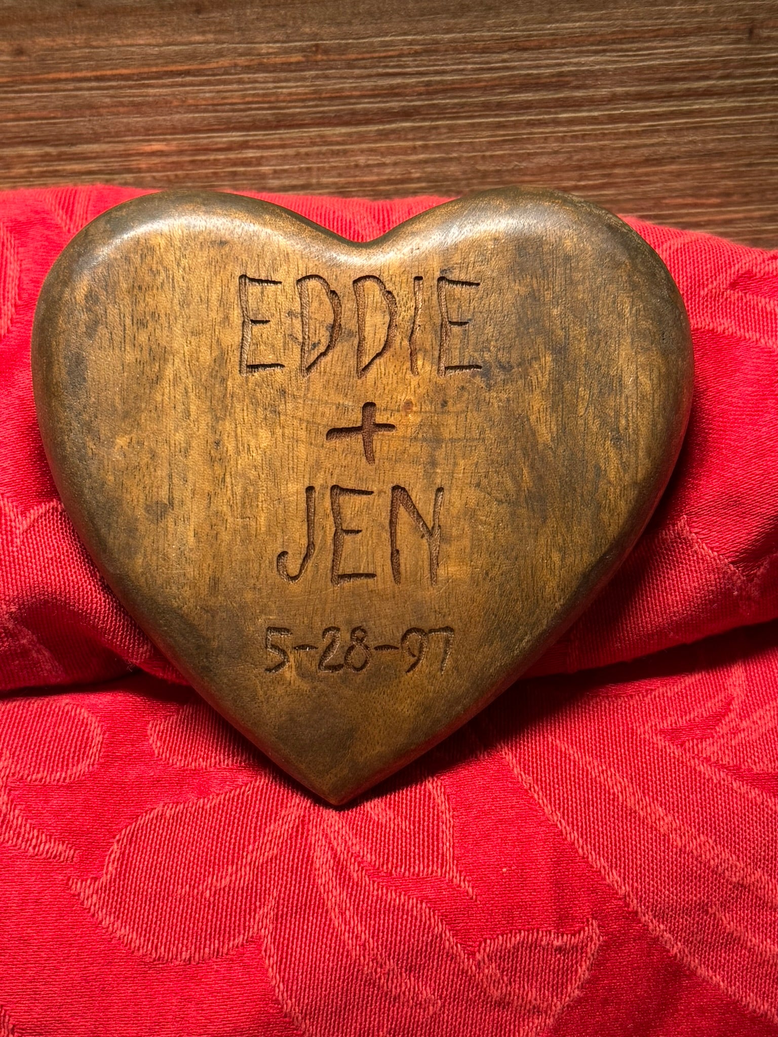 Rustic wooden heart engraved with the names 'Eddie + Jen' and the date '5-28-97,' displayed on a red fabric background.