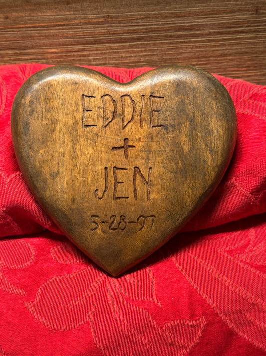 Rustic wooden heart engraved with the names 'Eddie + Jen' and the date '5-28-97,' displayed on a red fabric background.