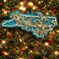 Teal North Carolina state ornament with laser-cut dogwood flower overlay, hanging on a Christmas tree