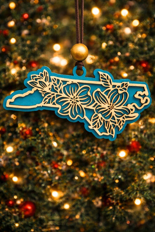 Teal North Carolina state ornament with laser-cut dogwood flower overlay, hanging on a Christmas tree