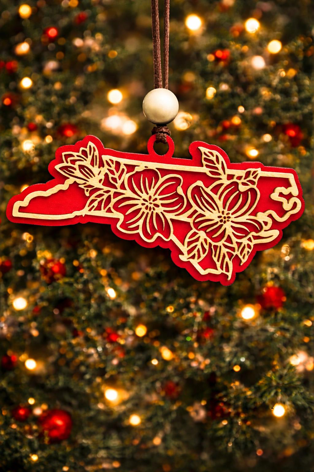 Red North Carolina state ornament with laser-cut dogwood flower overlay, hanging on a Christmas tree