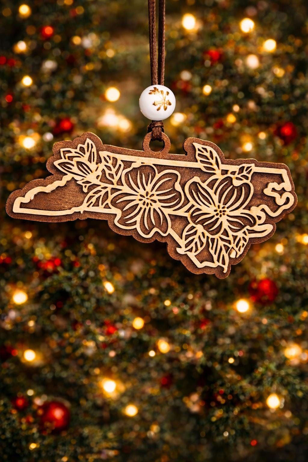 Walnut North Carolina state ornament with laser-cut dogwood flower overlay, hanging on a Christmas tree