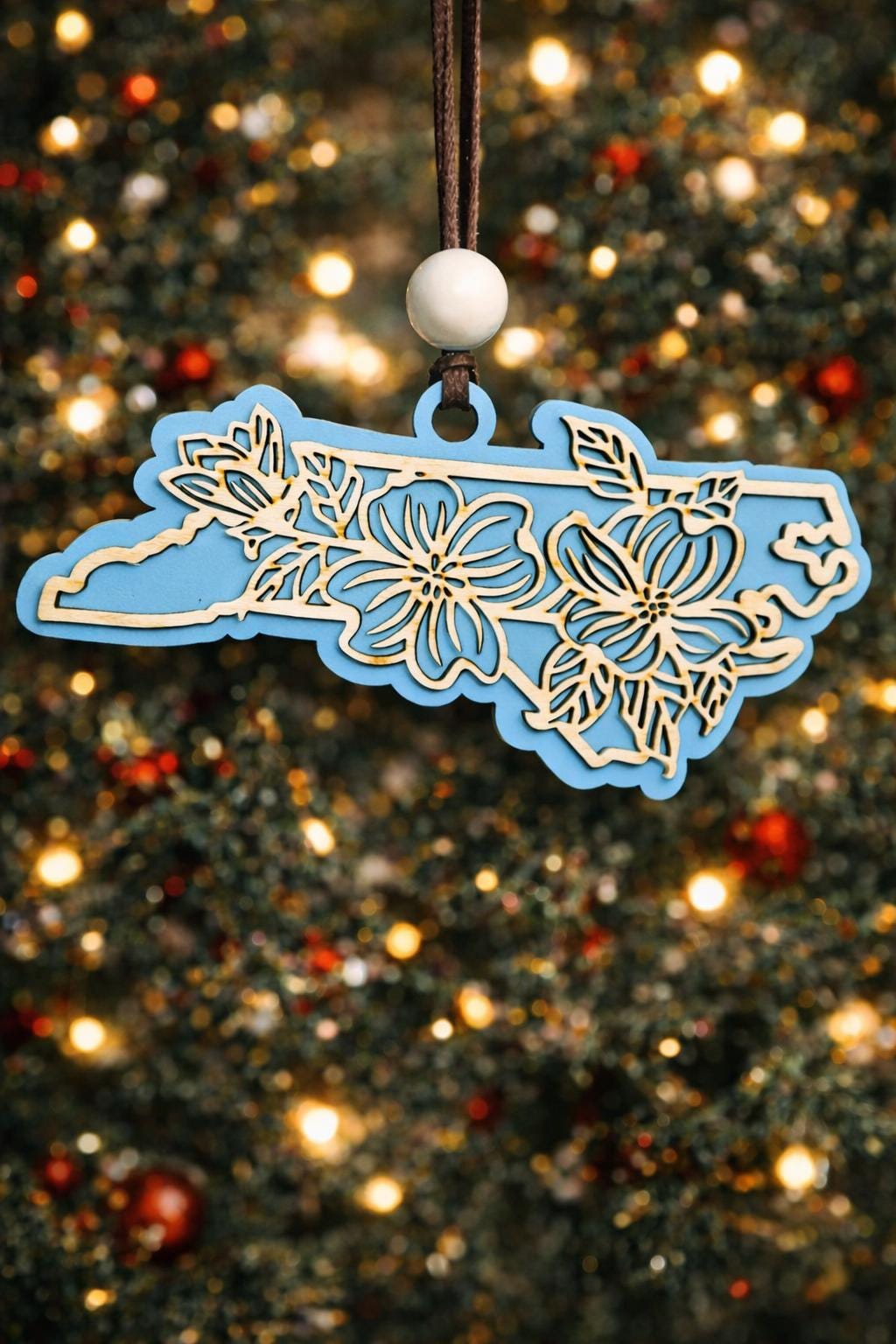 Sky Blue North Carolina state ornament with laser-cut dogwood flower overlay, hanging on a Christmas tree